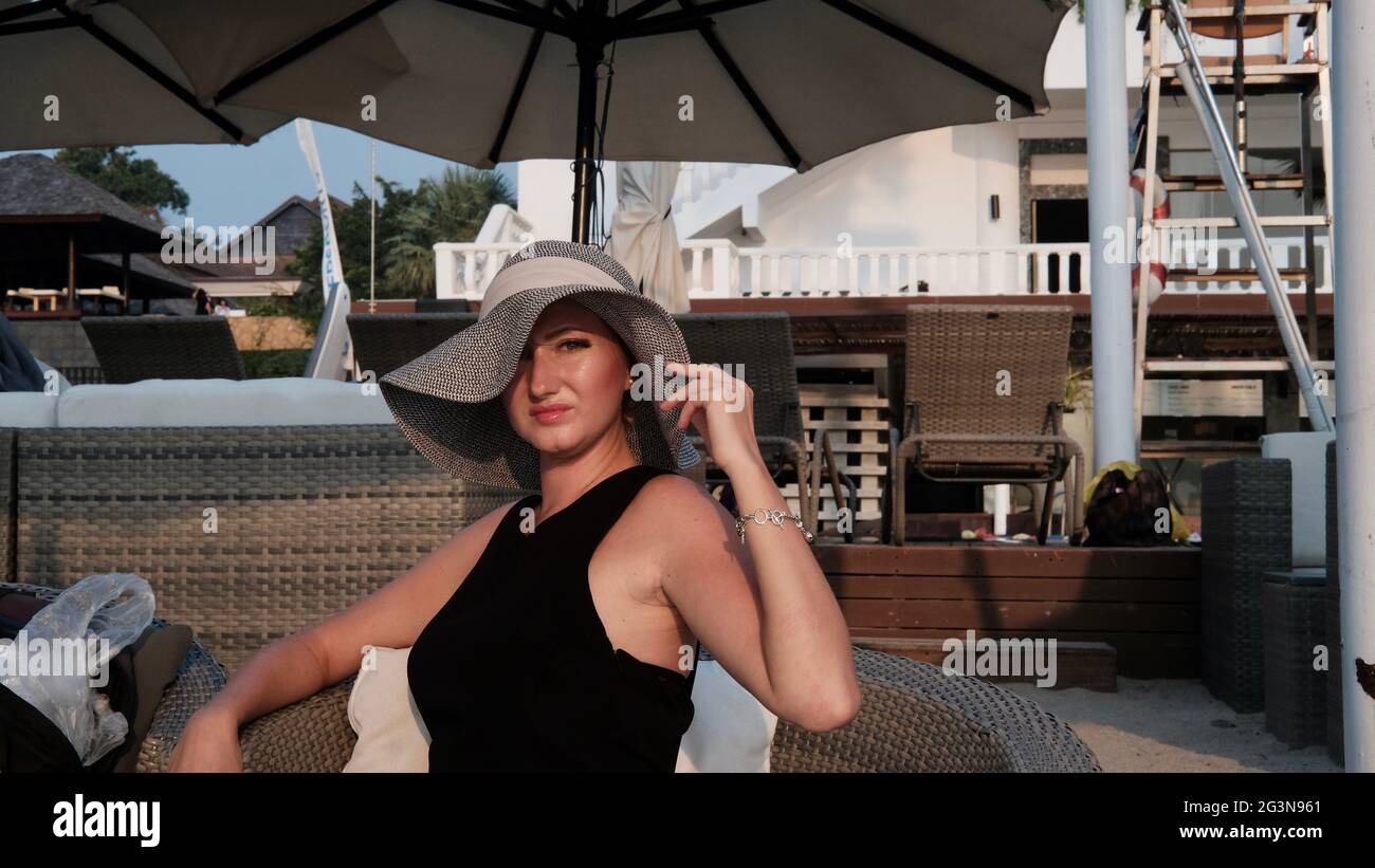 Osteuropäische kaukasische Frau mittleren Alters blondes Haar helle Haut blasse Teint in schwarzem Kleid grauen Strohhut außerhalb der Sommerzeit sitzen Stockfoto