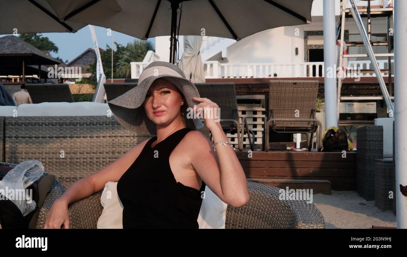 Osteuropäische kaukasische Frau mittleren Alters blondes Haar helle Haut blasse Teint in schwarzem Kleid grauen Strohhut außerhalb der Sommerzeit sitzen Stockfoto