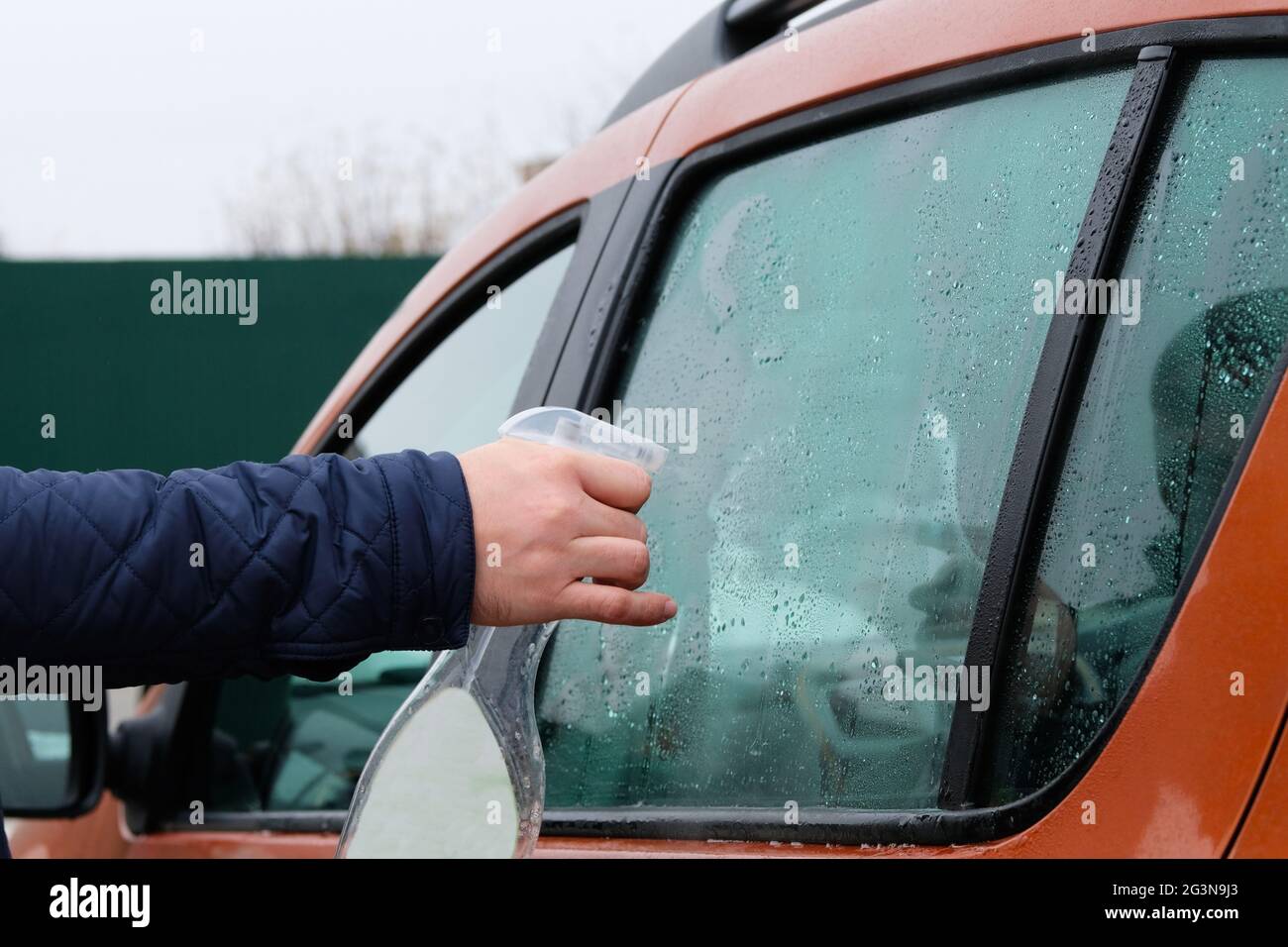Mann, der sein orangefarbenes Auto mit Spray säubert. Autofenster waschen. Tropfen auf Glas. Stockfoto