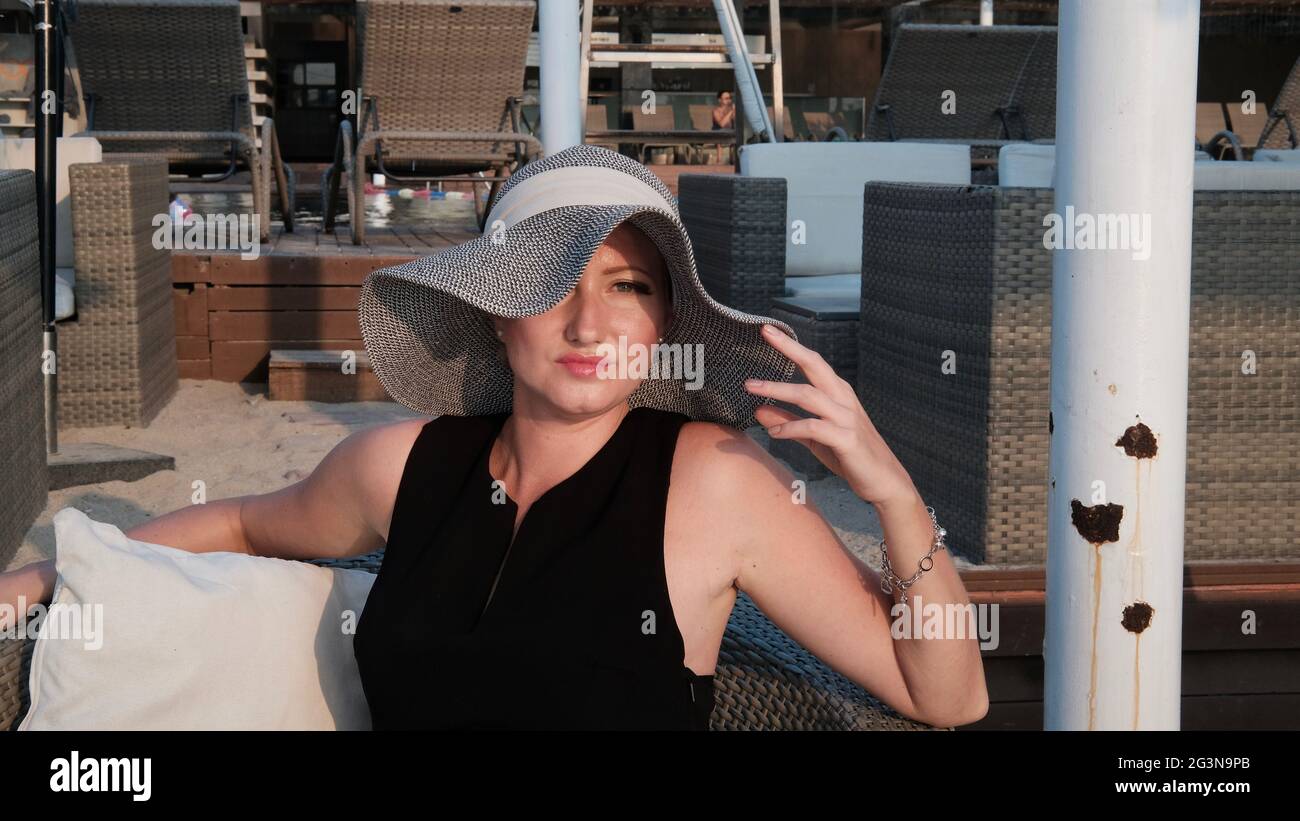 Osteuropäische kaukasische Frau mittleren Alters blondes Haar helle Haut blasse Teint in schwarzem Kleid grauen Strohhut außerhalb der Sommerzeit sitzen Stockfoto