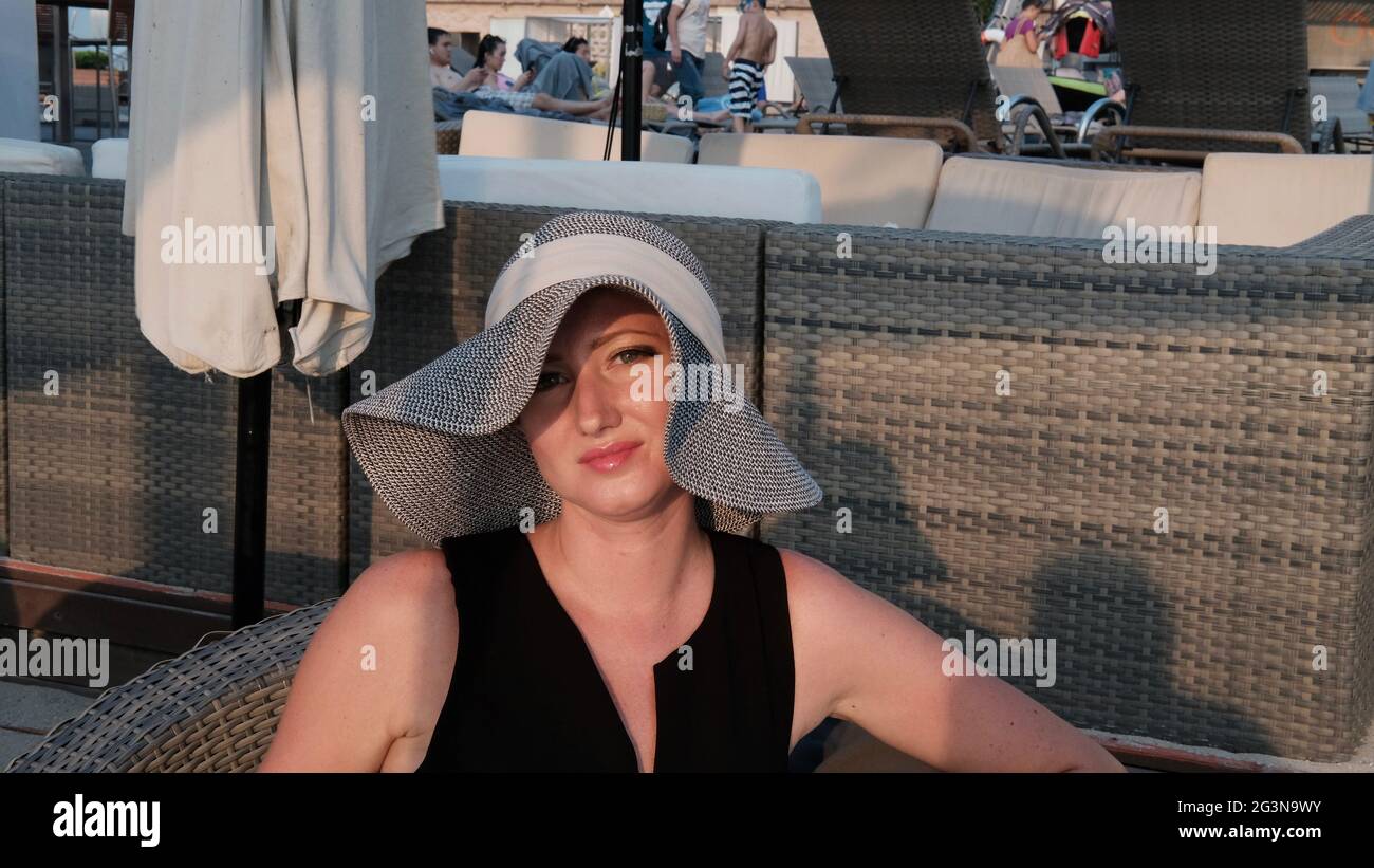Osteuropäische kaukasische Frau mittleren Alters blondes Haar helle Haut blasse Teint in schwarzem Kleid grauen Strohhut außerhalb der Sommerzeit sitzen Stockfoto