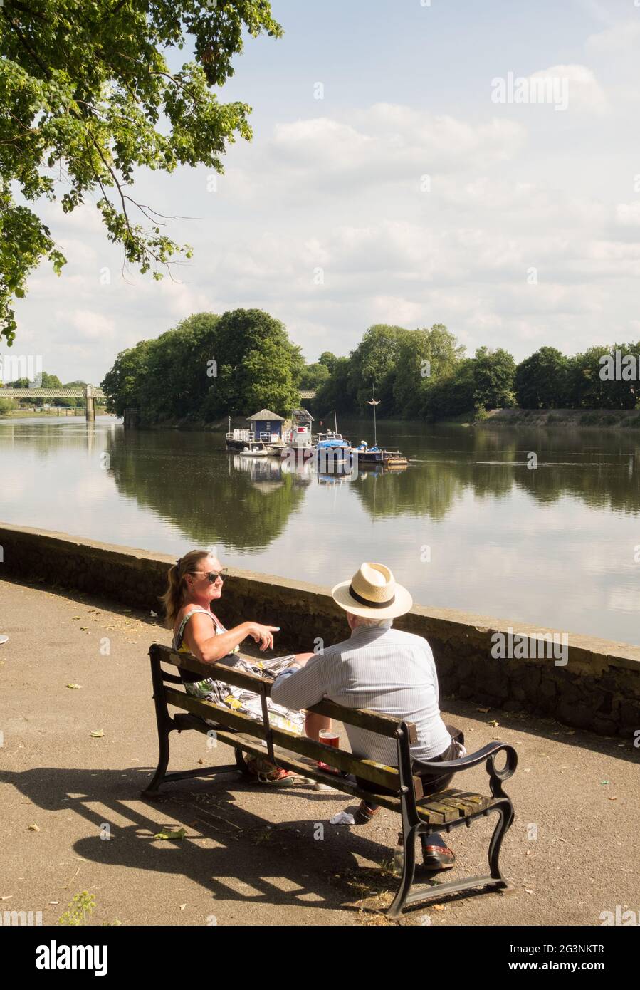 Ein Pärchen, das sich unterhielt und auf einer Parkbank neben der Themse saß, aus der Sicht von Strand-on-the-Green, Chiswick, Hounslow, London, England, GROSSBRITANNIEN Stockfoto
