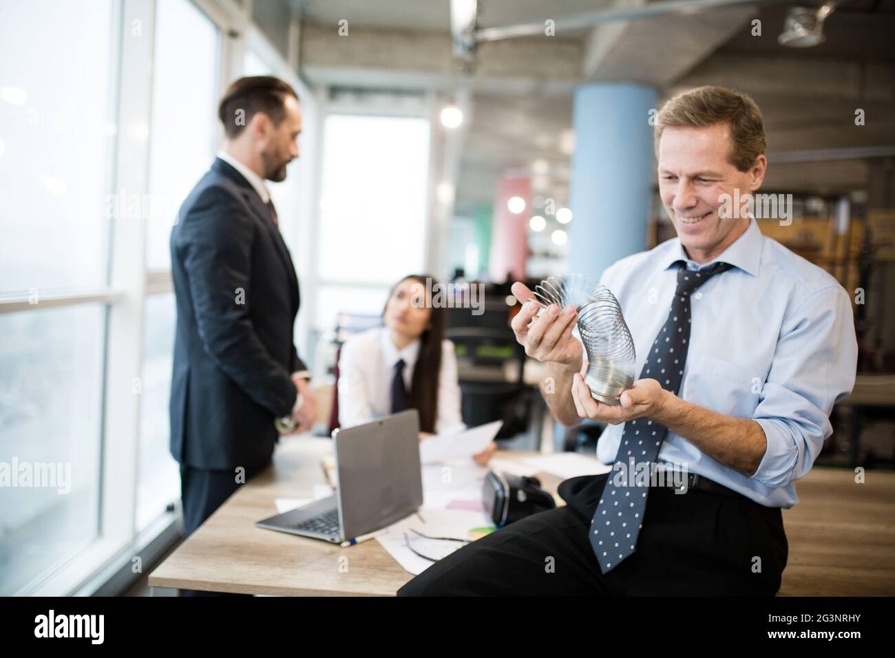 Ein Geschäftsmann spielt mit einem Metallspielzeug, das slinky ist. Büroangestellte sprechen im Büro dahinter Stockfoto