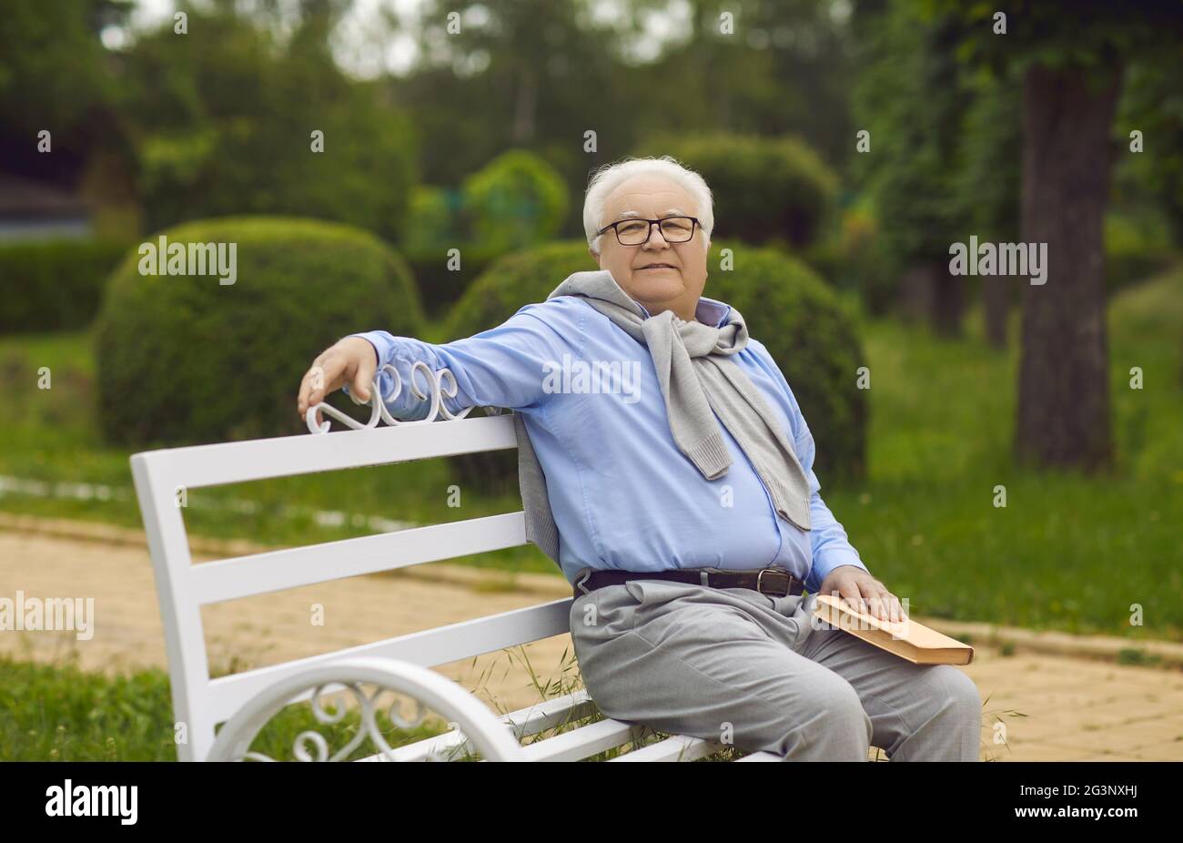 Ein älterer Mann sitzt auf einer Bank und liest ein Buch. Ein älterer Mann mit weißem Haar ruht auf einer Parkbank Stockfoto