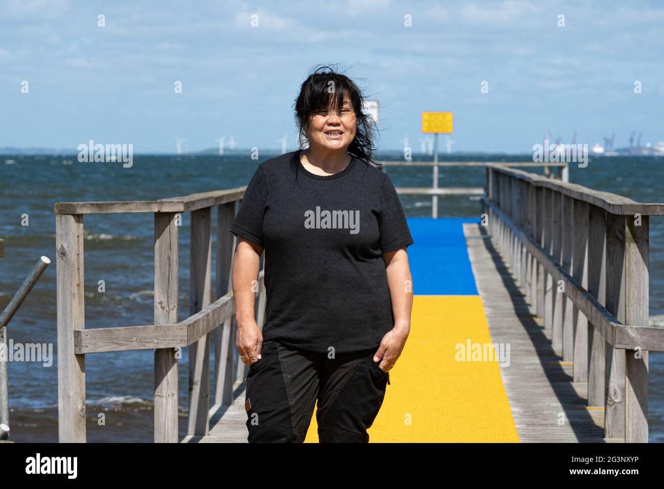 Eine asiatische Frau mittleren Alters auf einem Steg mit einem gelben und blauen Teppich und einem blauen Ozean und Himmel im Hintergrund. Bild aus der Grafschaft Scania, Schweden Stockfoto