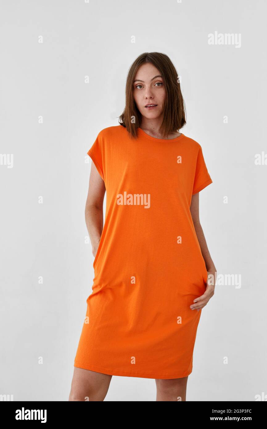 Niedliches weibliches Modell in legerem orangefarbenem Kleid mit Blick auf die Kamera vor weißem Hintergrund Stockfoto