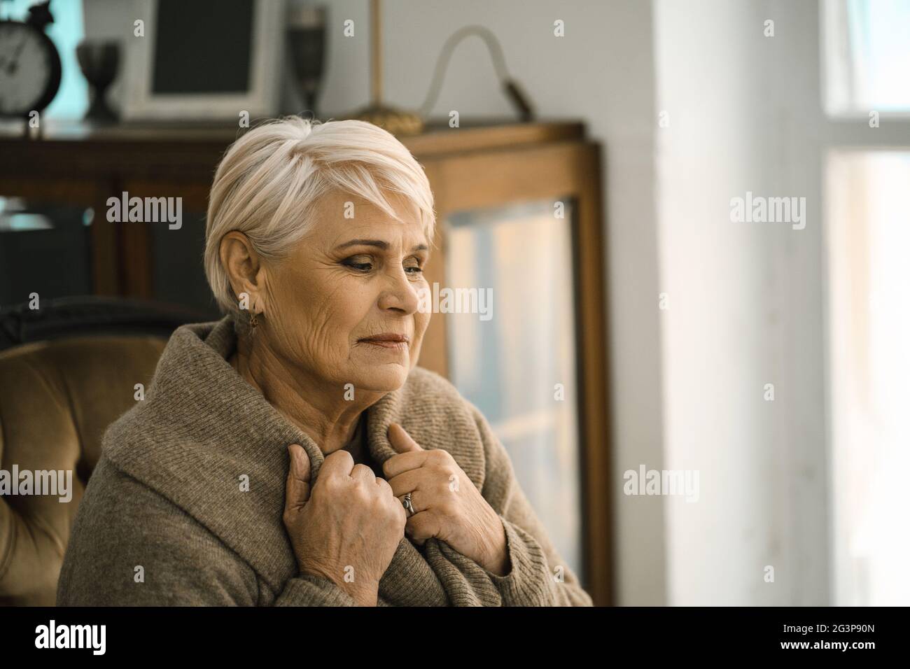 Nachdenkliche ältere Frau, die sich in EINEN warmen Pullover einwickelt, der auf dem gemütlichen Sofa siegt Stockfoto