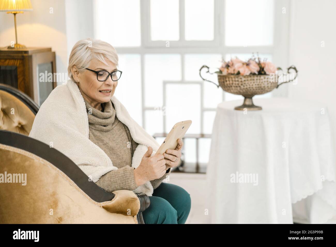 Elegante Ältere Frau In Wollpullover Liest Etwas Auf Ihrem Tablet Und Lächelt Stockfoto