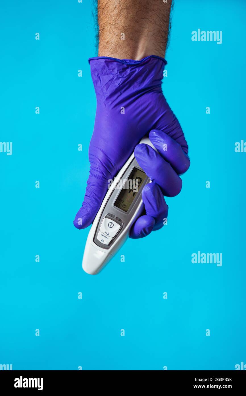 Die Hand eines Arztes trägt einen violetten Handschuh und hält ein Thermometer auf blauem Hintergrund. Corona-Virenschutz. Stockfoto
