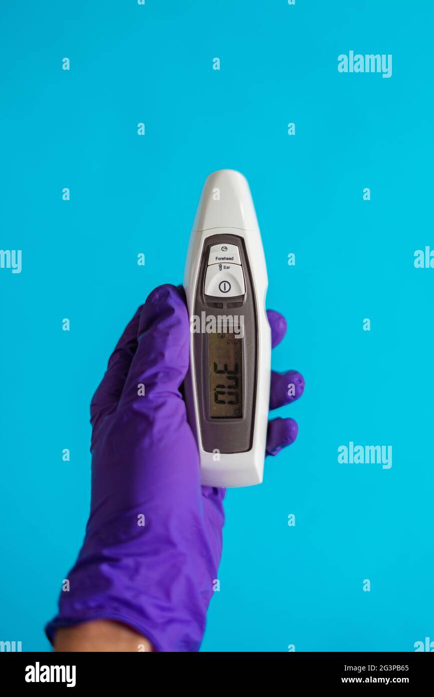Die Hand eines Arztes trägt einen violetten Handschuh und hält ein Thermometer auf blauem Hintergrund. Corona-Virenschutz. (Ansicht von oben) Stockfoto