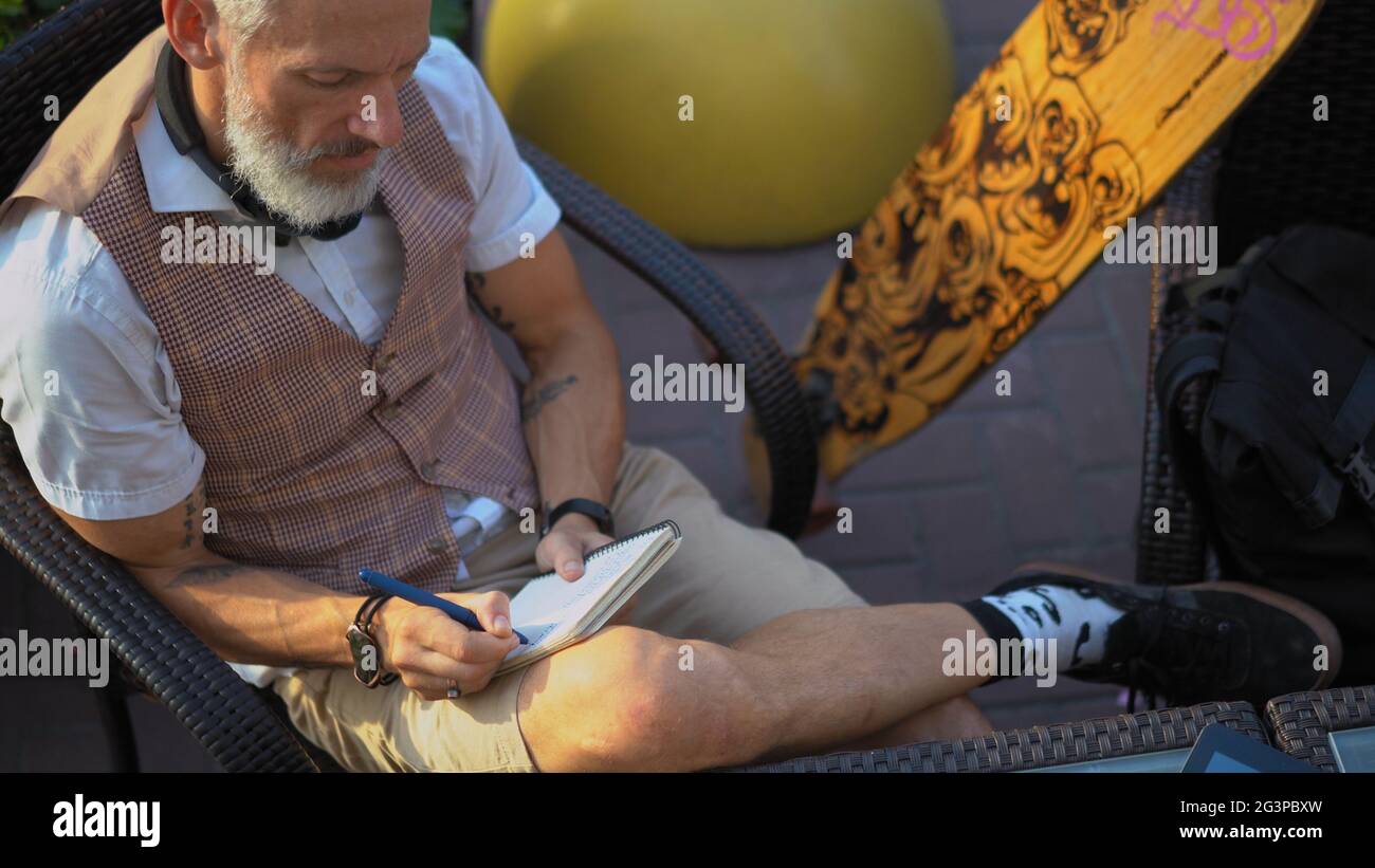Ein intelligenter Mann schreibt in einem Straßencafé in ein Notizbuch und ruht sich nach einem Longboard aus Stockfoto