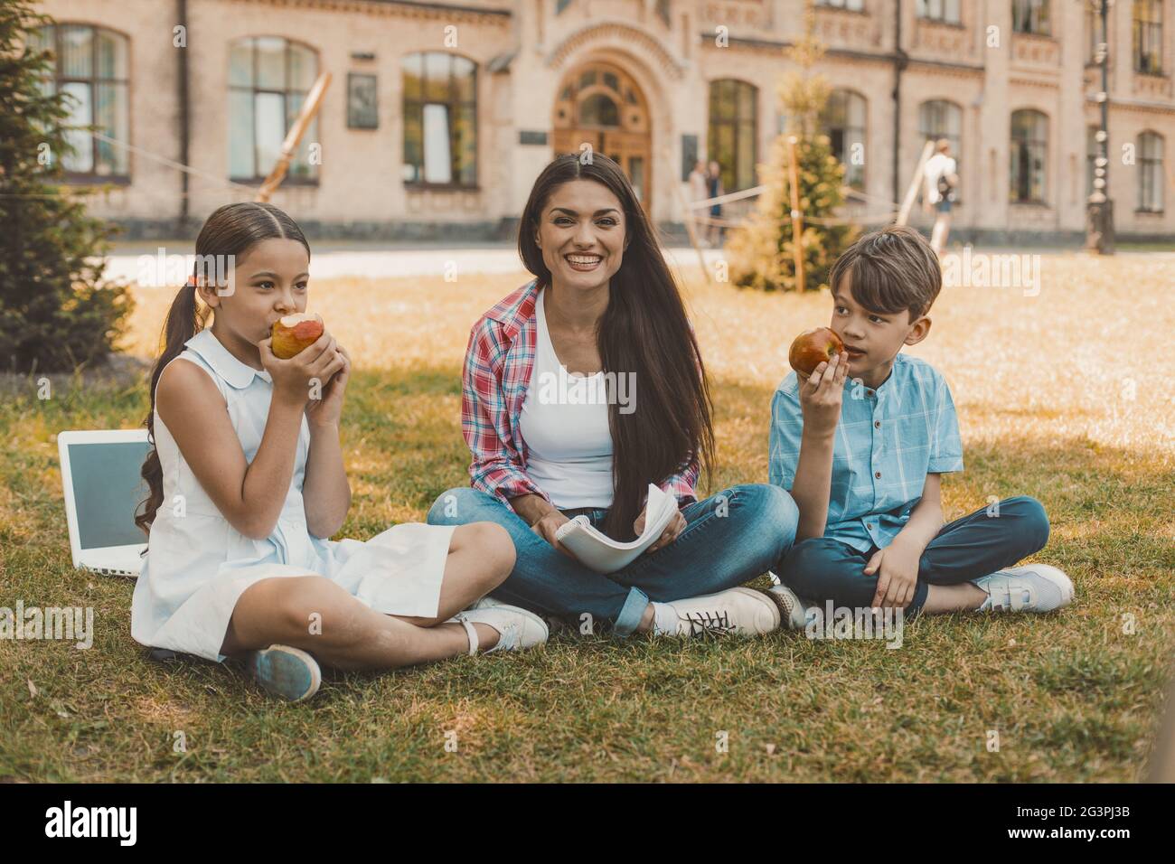 Junge und Mädchen essen Äpfel, während junge Frau auf Gras sitzt Stockfoto