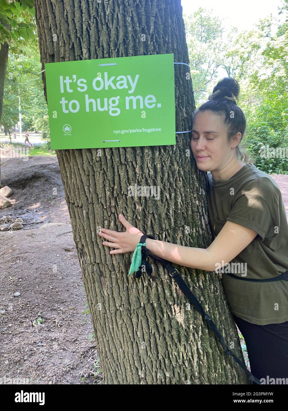 Die junge Frau umarmt einen Baum im Prospect Park, der hier und dort Schilder angebracht hat und die Leute daran erinnert, einen Baum zu umarmen. Die Nähe zur Natur kann Stress lindern, den Blutdruck senken und Depressionen lindern. Stockfoto