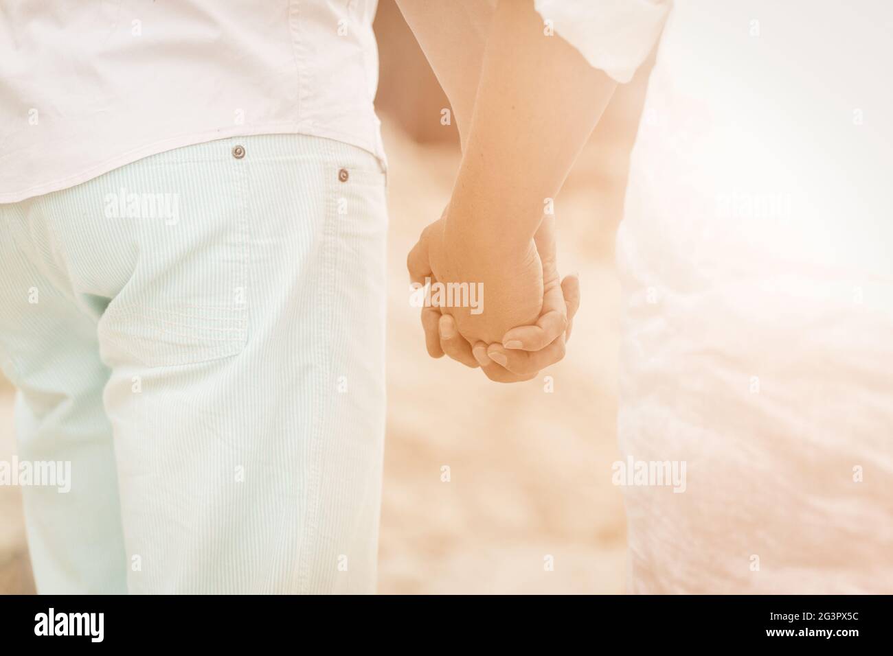 Paar hält sich am Meer die Hände. Rückansicht Mann und Frau in hellen Kleidern, die Hände halten, gehen zusammen am Strand entlang auf einer Sonne Stockfoto