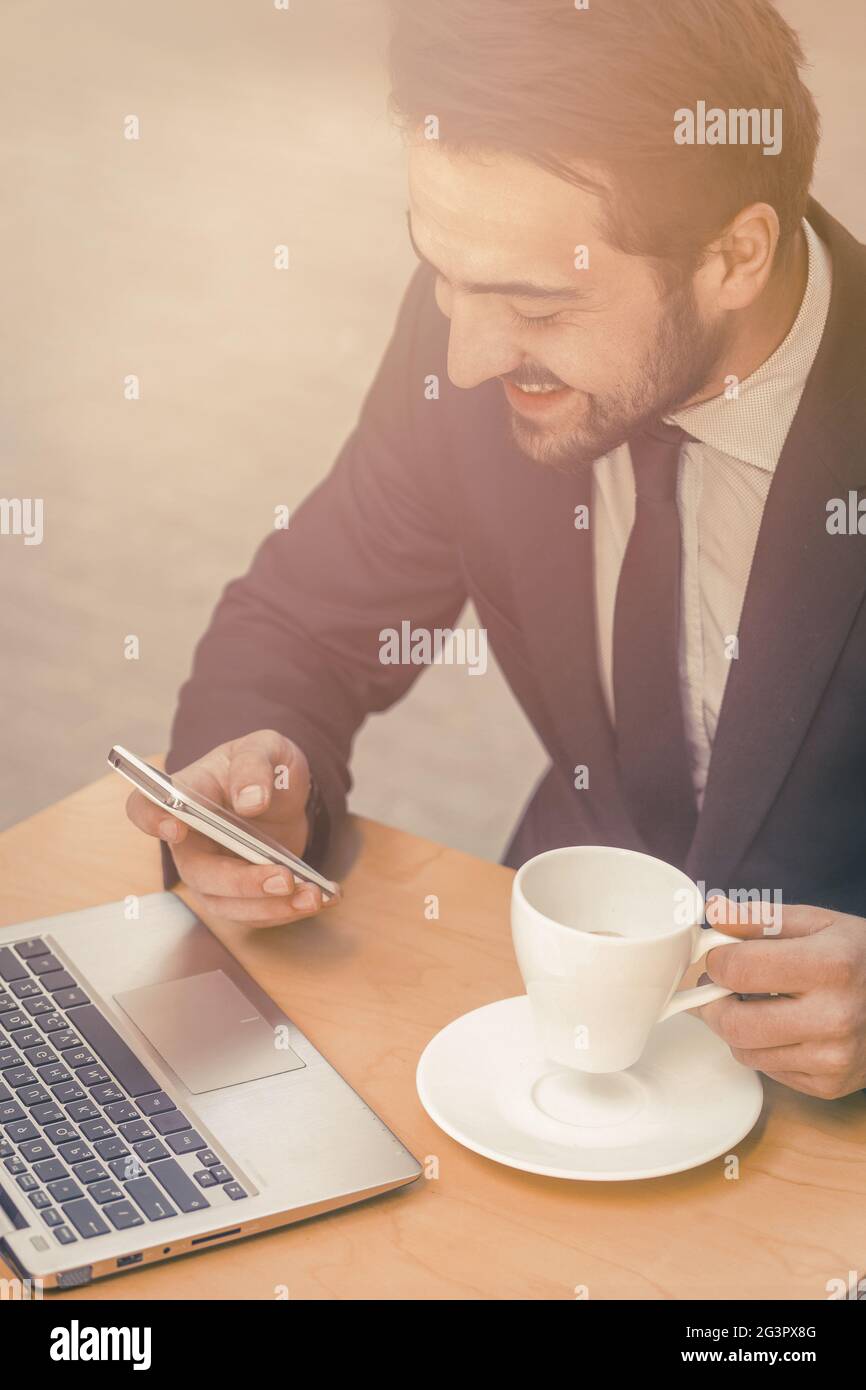 Glücklicher Geschäftsmann beim Lesen von Nachrichten auf dem Handy. Lachender Mann trinkt Kaffee im Straßencafé und plaudert auf dem Smartphone, während w Stockfoto