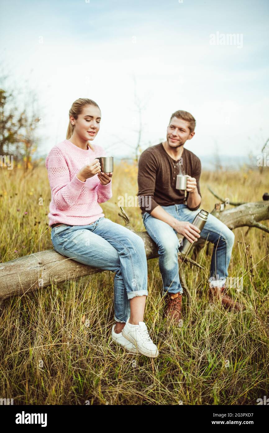 Tee-Party oder Kaffee trinken im Freien. Ein fröhliches Paar trinkt heißen Kaffee oder Tee und kommuniziert auf einem Holzbalken im Freien Stockfoto