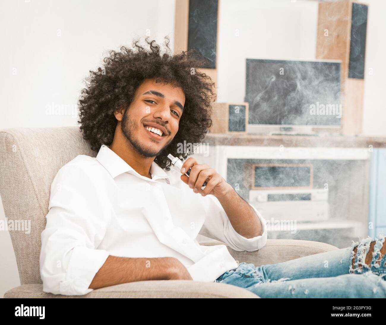 Junger freiberuflicher Mann entspannt sich beim Rauchen von E-Zigarette. Shaggy Macho lächelt charmant, während sie auf die Kamera schaut. Getöntes Bild Stockfoto