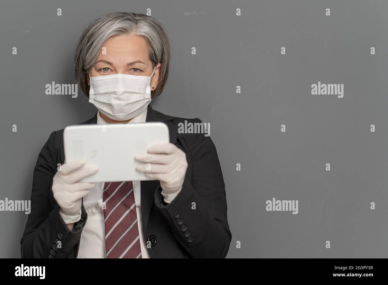 Grauhaarige Geschäftsfrau arbeitet Notizblock. Kaukasische Dame hält Tablet Blick auf die Kamera auf grauem Hintergrund mit Textraum auf rechts Stockfoto