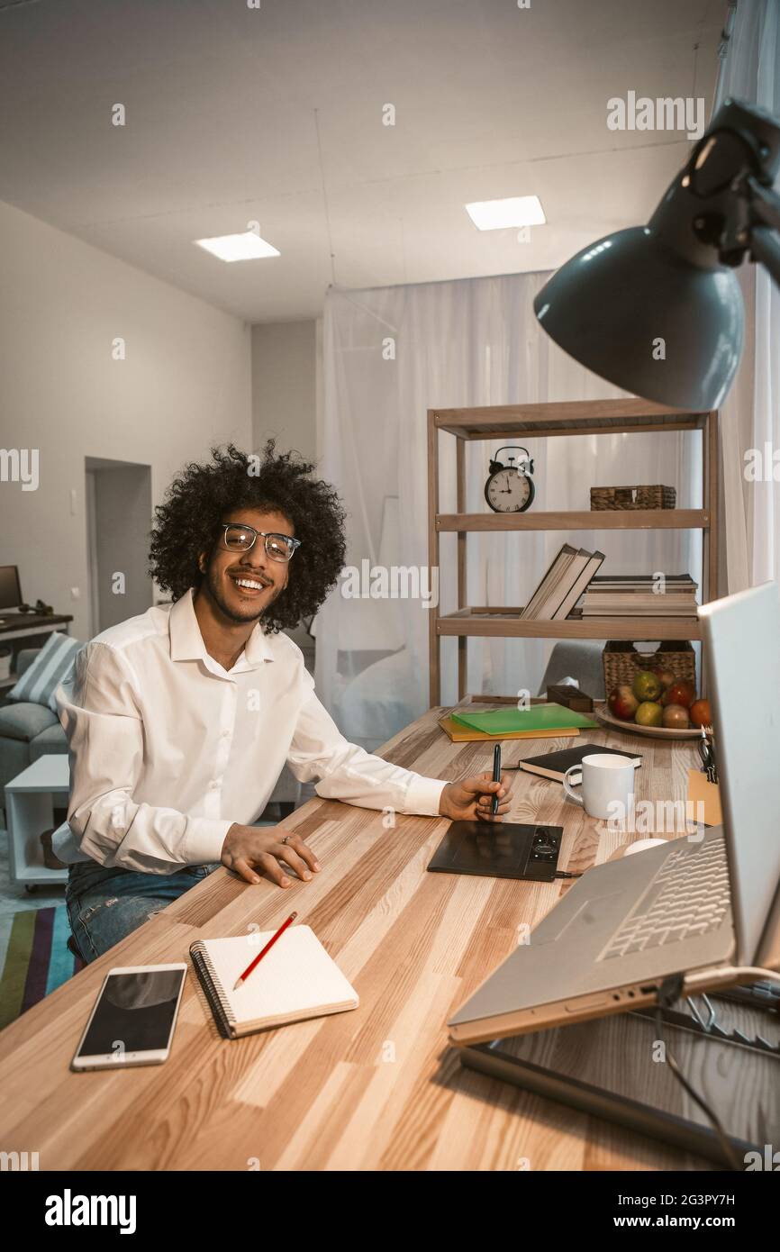 Kreativer Mann, der gerne von zu Hause aus arbeitet. Der smarte arabische Kerl lächelt am Schreibtisch mit einem grafischen Tablet und einem Computer auf dem Schreibtisch in gemütlicher Atmosphäre Stockfoto