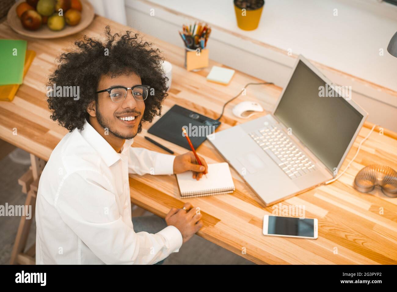 Intellectual Millennial schreibt in einem Notizbuch, das die Kamera anschaut, während er am Arbeitsplatz in einem gemütlichen, gemütlichen Interieur sitzt. Copywrite Stockfoto