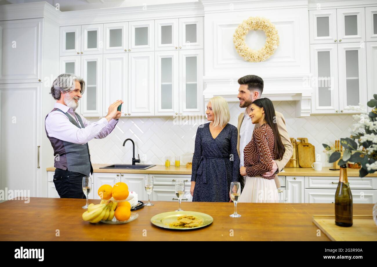 Eleganter älterer grauhaariger Mann Großvater, der während der Geburtstagsfeier seine Frau und sein junges Paar fotografiert, die in einer stilvollen Küche zu Hause mit Obst, Süßigkeiten und Champagner auf dem Tisch stattfindet Stockfoto