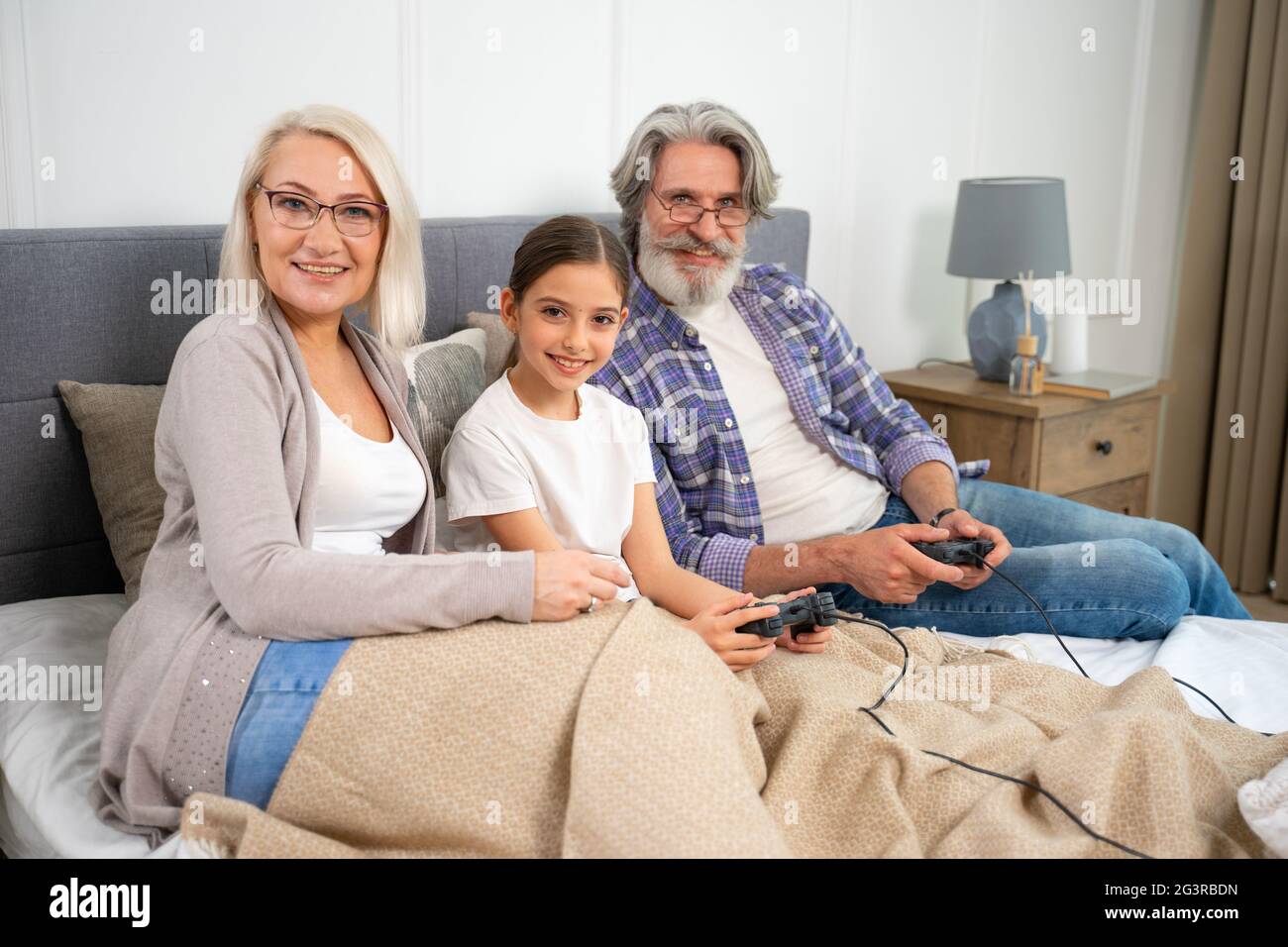 Porträt der entzückenden kleinen Enkelin und älteren Großeltern lächeln vor der Kamera, während sie gemeinsam Videospiele spielen, auf dem Bett sitzen und am Wochenende zu Hause die Freizeit der Familie genießen Stockfoto