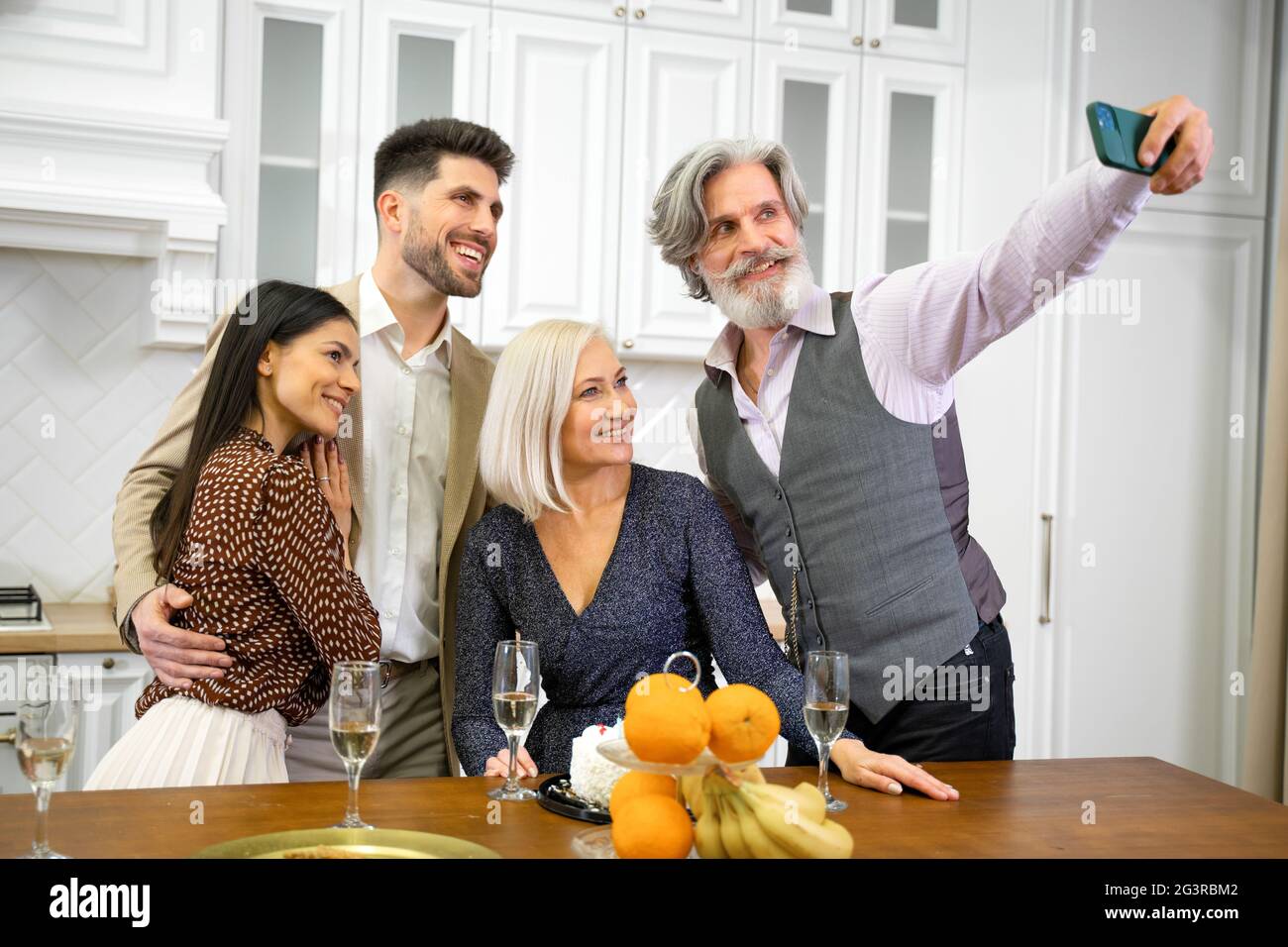 Positive Großeltern und junge Paare in formeller Kleidung, die Selfie machen oder Videoanruf auf dem Smartphone machen, die Kamera betrachten und während der Feiertagsfeier mit der Familie zu Hause lächeln Stockfoto