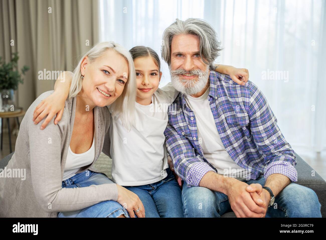 Porträt einer niedlichen kleinen Enkelin und ihrer im Ruhestand lächelnden schönen Großeltern, die im Wohnzimmer zu Hause auf dem Sofa sitzen und Zeit miteinander verbringen und besondere Momente mit der Familie teilen Stockfoto