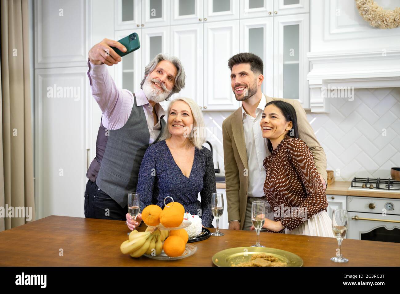 Glückliche Großeltern der Familie und ein junges lächelndes Paar in formeller festlicher Kleidung, das Selfie auf dem Smartphone macht, die Kamera anschaut und während der Geburtstagsfeier in der Küche zu Hause lächelt Stockfoto