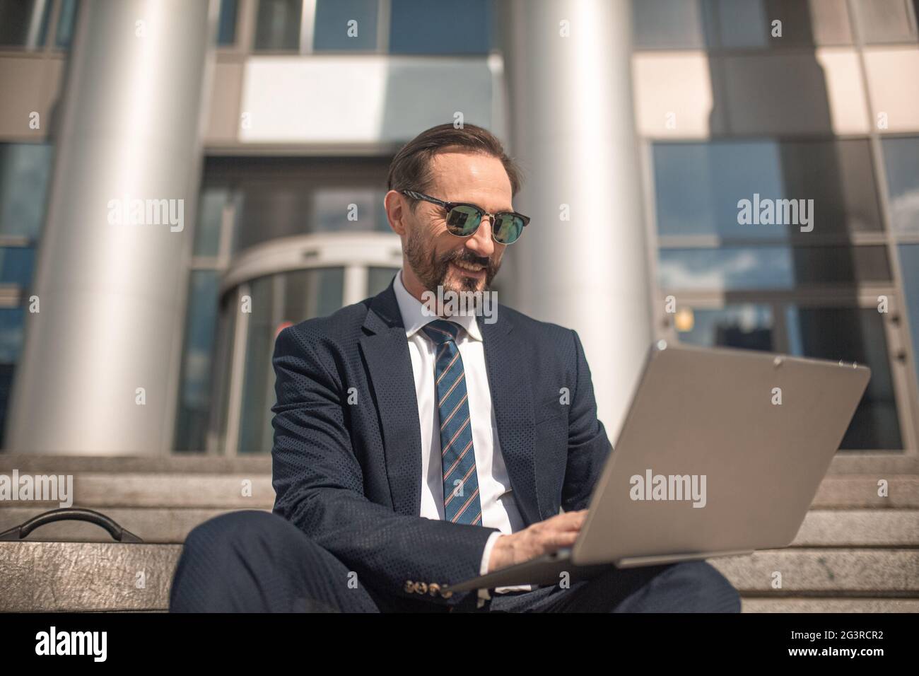 Ein erfolgreicher Geschäftsmann arbeitet auf einem Laptop auf der Treppe des Business Centers. Lächelnder Mann mit Business-Anzug und Sonnenbrille. Hi Stockfoto