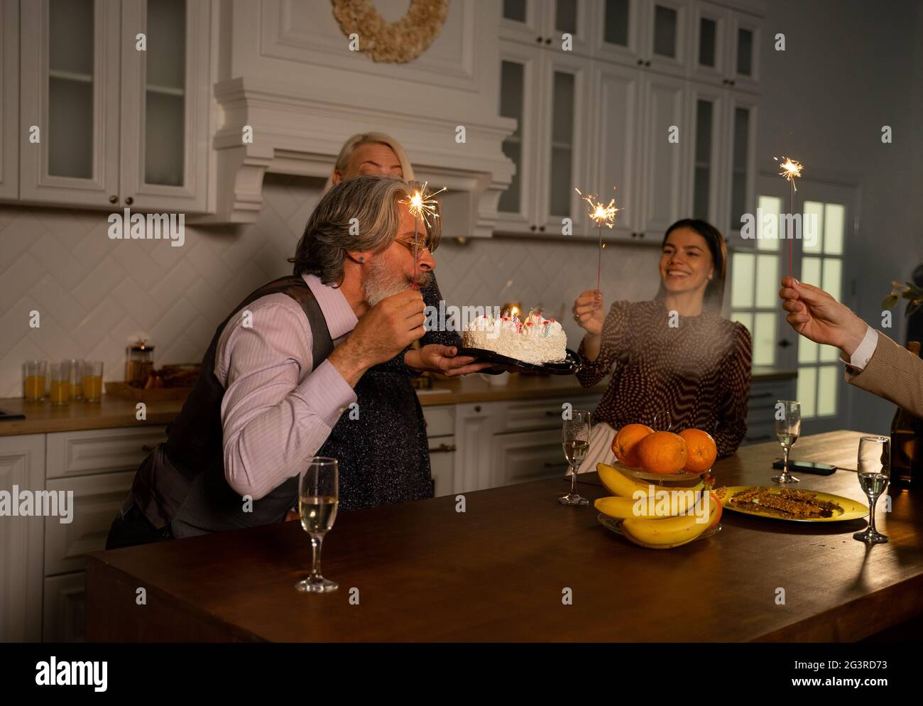 Großvater bläst Kerzen auf Geburtstagstorte aus und wünscht sich, während er zu Hause hinter dem Küchentisch sitzt, seine Frau Kuchen hält und Gäste Wunderkerzen hält. Konzept für Familienfeiern Stockfoto