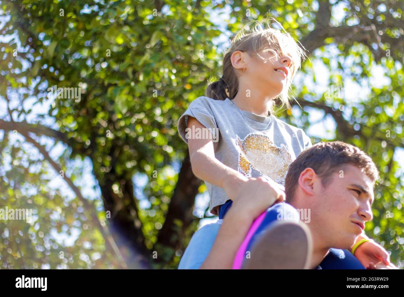 Defocused Bruder Reiten Schwester auf dem Rücken. Portrait von glücklichen Mädchen auf Mann Schultern, Huckepack. Mädchen fliegen. Familie spielt draußen. Grüner Baum verschwimmt Stockfoto