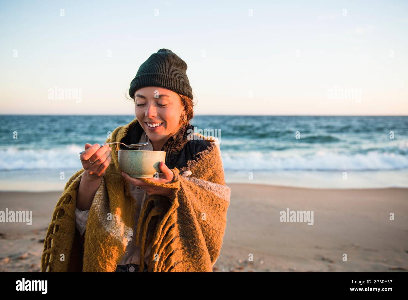 Junge Frau genießt eine Schüssel Suppe, während sie im Herbst am Strand campen Stockfoto