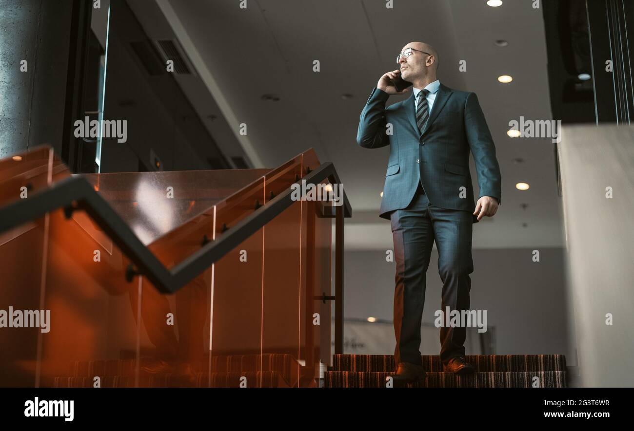 Geschäftsmann, der im Business Center über eine Treppe steht und mit dem Handy telefoniert. Schöner kaukasischer Mann, der sich das Copy Spa ansieht Stockfoto