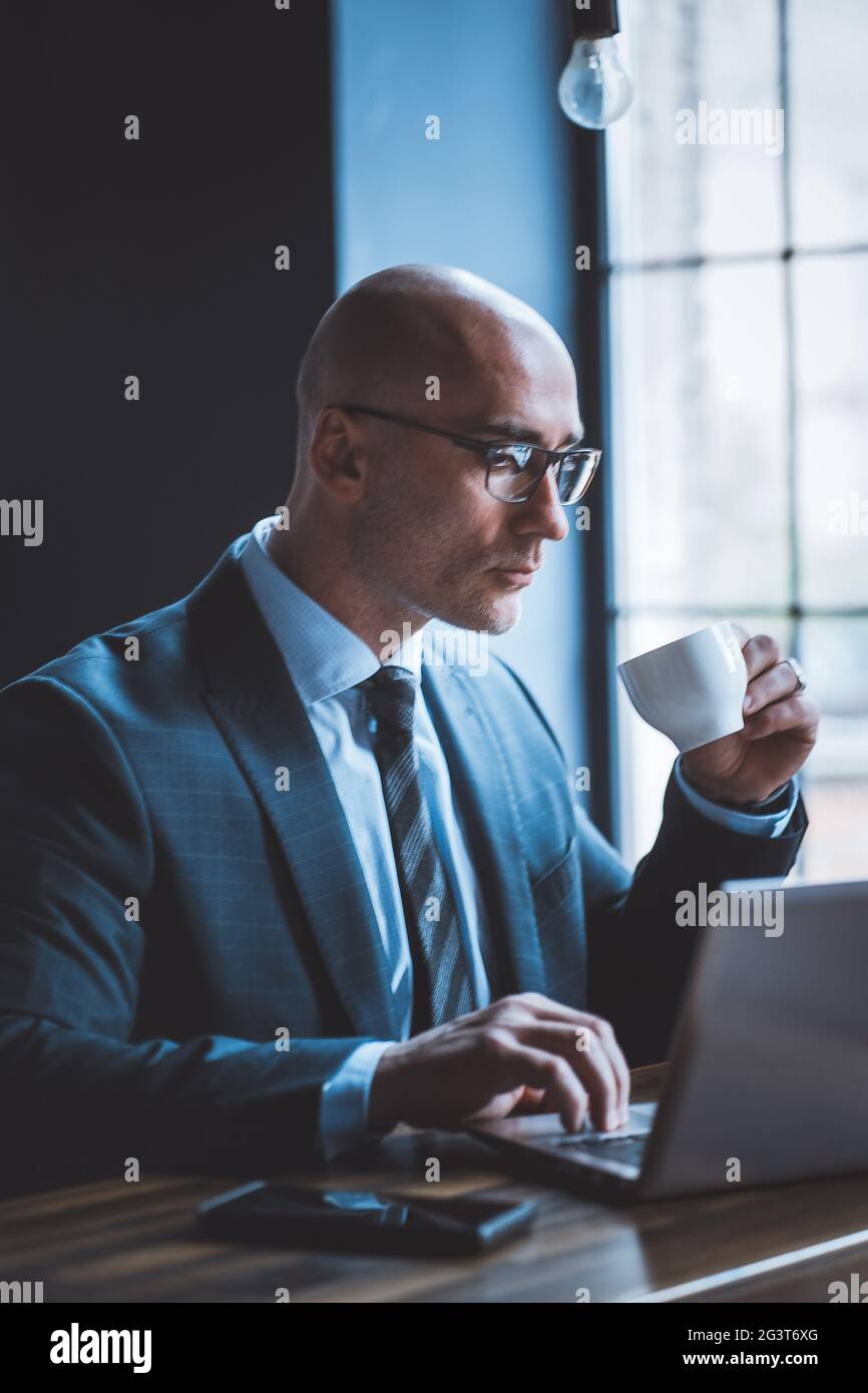 Ein respektabler Geschäftsmann trinkt Kaffee und arbeitet mit einem Computer vor dem Fenster. Seitenansicht des gut aussehenden kaukasischen schönen Typi Stockfoto