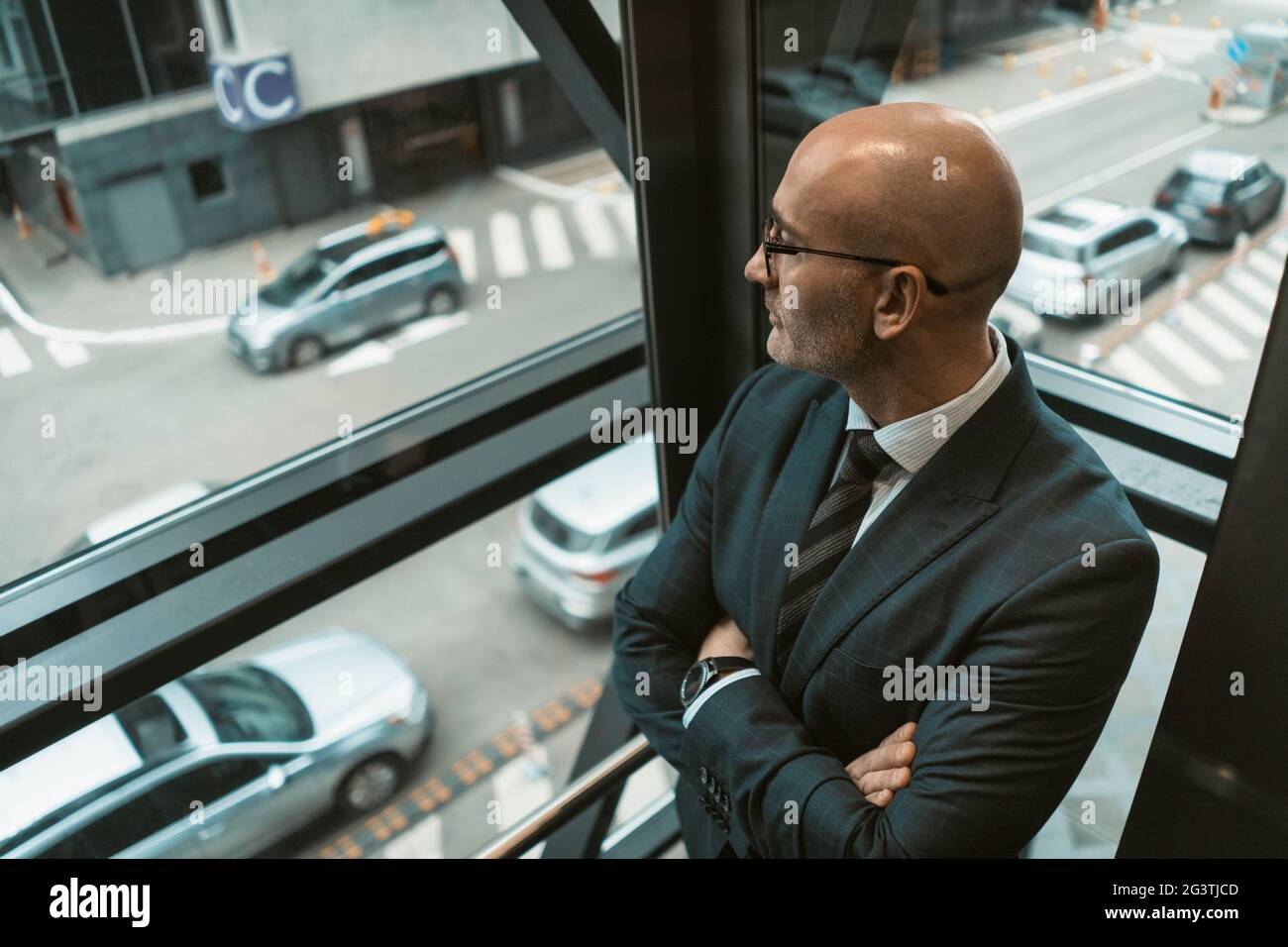 Ansicht von oben. Kontemplativer kahler Geschäftsmann in Anzug und Brille, der mit einem geschäftigen Treiben im Fenster oder Glasaufzug wegschaut Stockfoto