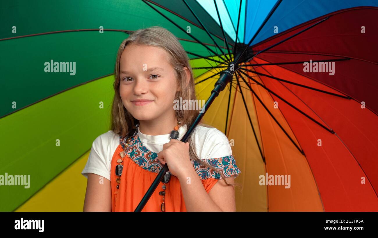 Kleines Mädchen in orangefarbenem Kleid mit Regenbogenfarben Regenschirm, das im Studio jubelt und die Kamera auf da isoliert betrachtet Stockfoto