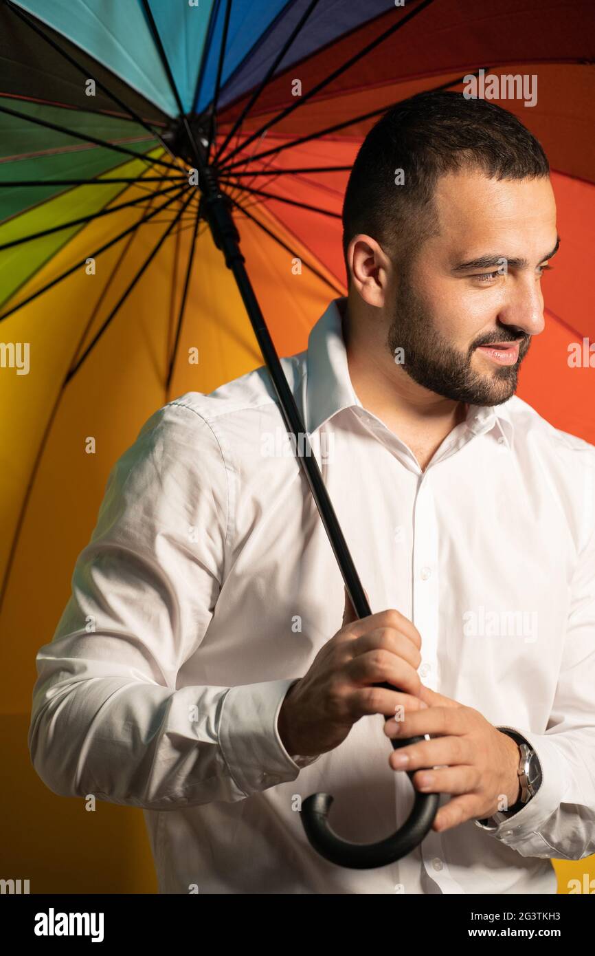Gutaussehender Kerl zur Unterstützung der LGBT-Gesellschaft. Nahaufnahme hübscher Bartjunge in weißem Hemd, der mit einem Regenbogenschirm hinter seinem Schirm steht Stockfoto