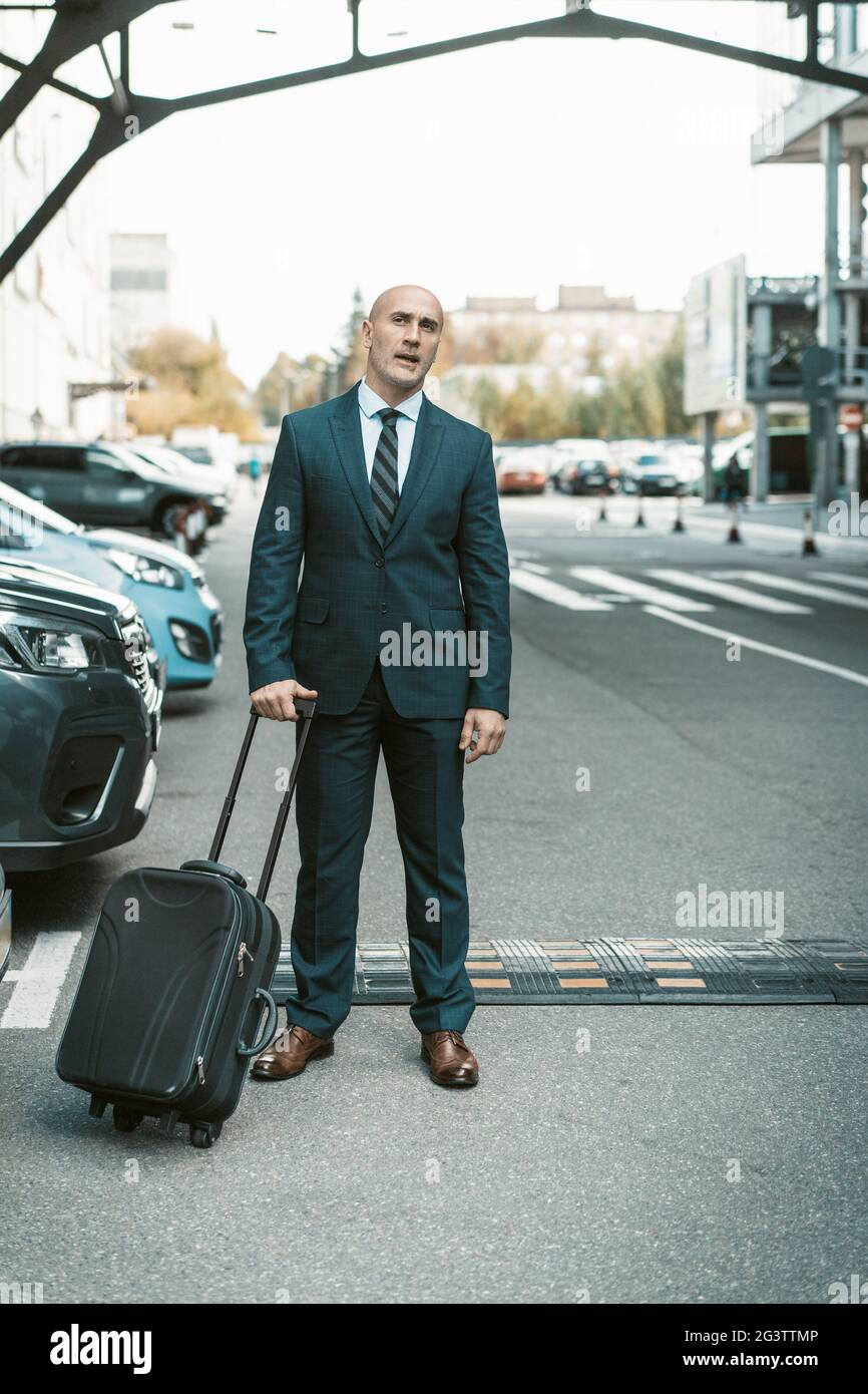 Warten auf seinen Fahrer oder sein Taxi schöner Geschäftsmann steht auf dem Parkplatz mit Koffer. Geschäftsmann in Anzug und Koffer Stockfoto