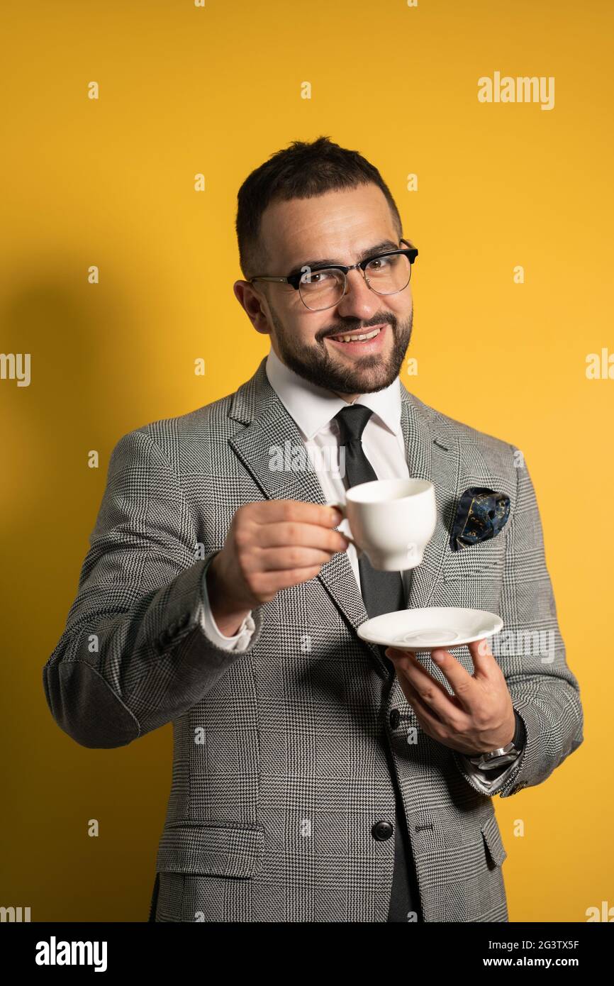 Fröhliche junge gut aussehende bärtige Mann in Augenbrille in formell Tragen Sie eine Tasse Kaffee auf gelbem Hintergrund isoliert halten Stockfoto