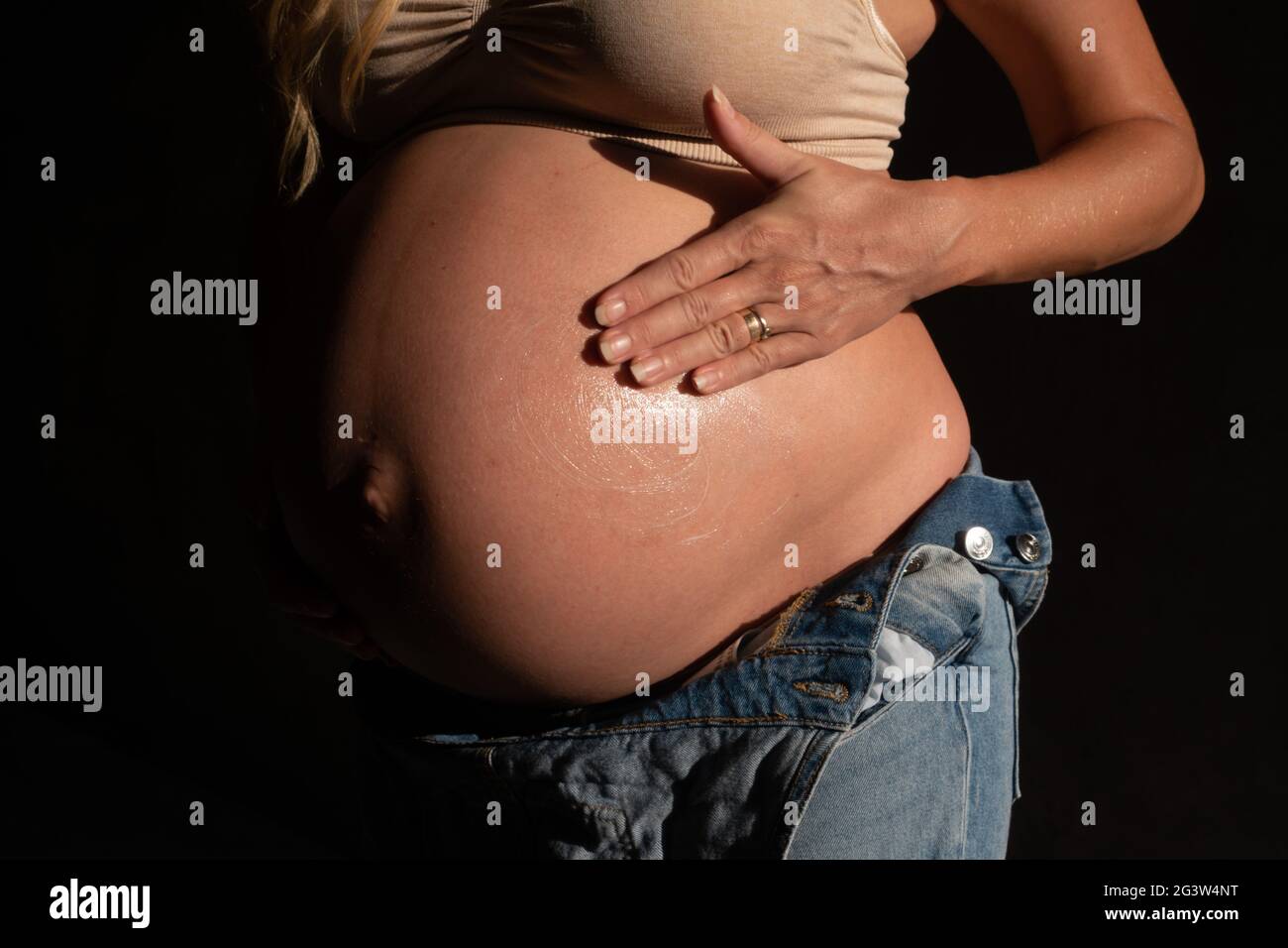 Beschnittenes Bild einer Schwangeren im Jeanssanzug und BH, die Lotion auf ihren Bauch auftragen Stockfoto