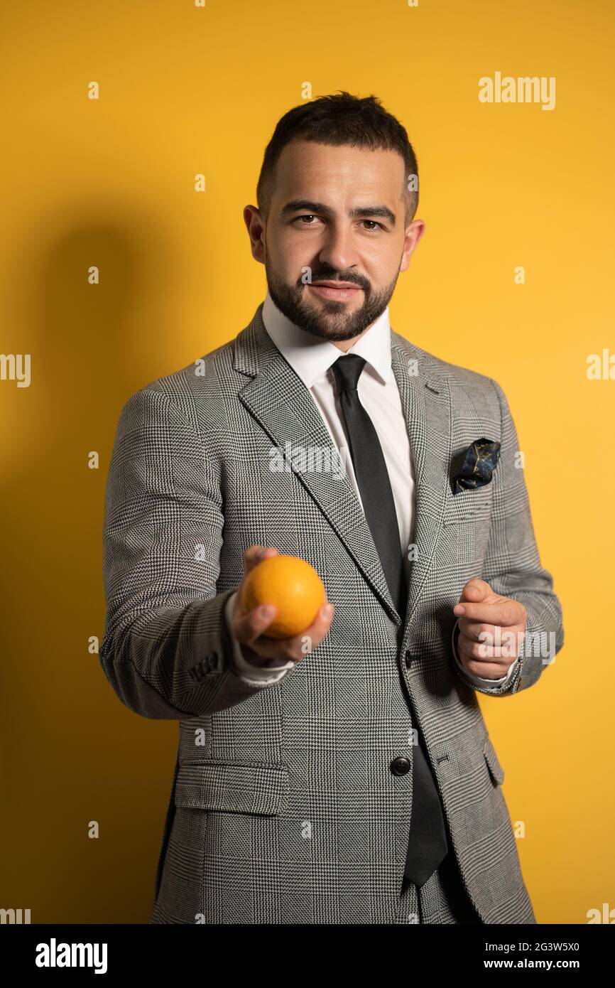 Hübscher eleganter Mann in grauem Anzug mit einer Brille hält seine Hand mit Orange in einer Hand aus, die isoliert auf Gelb steht Stockfoto