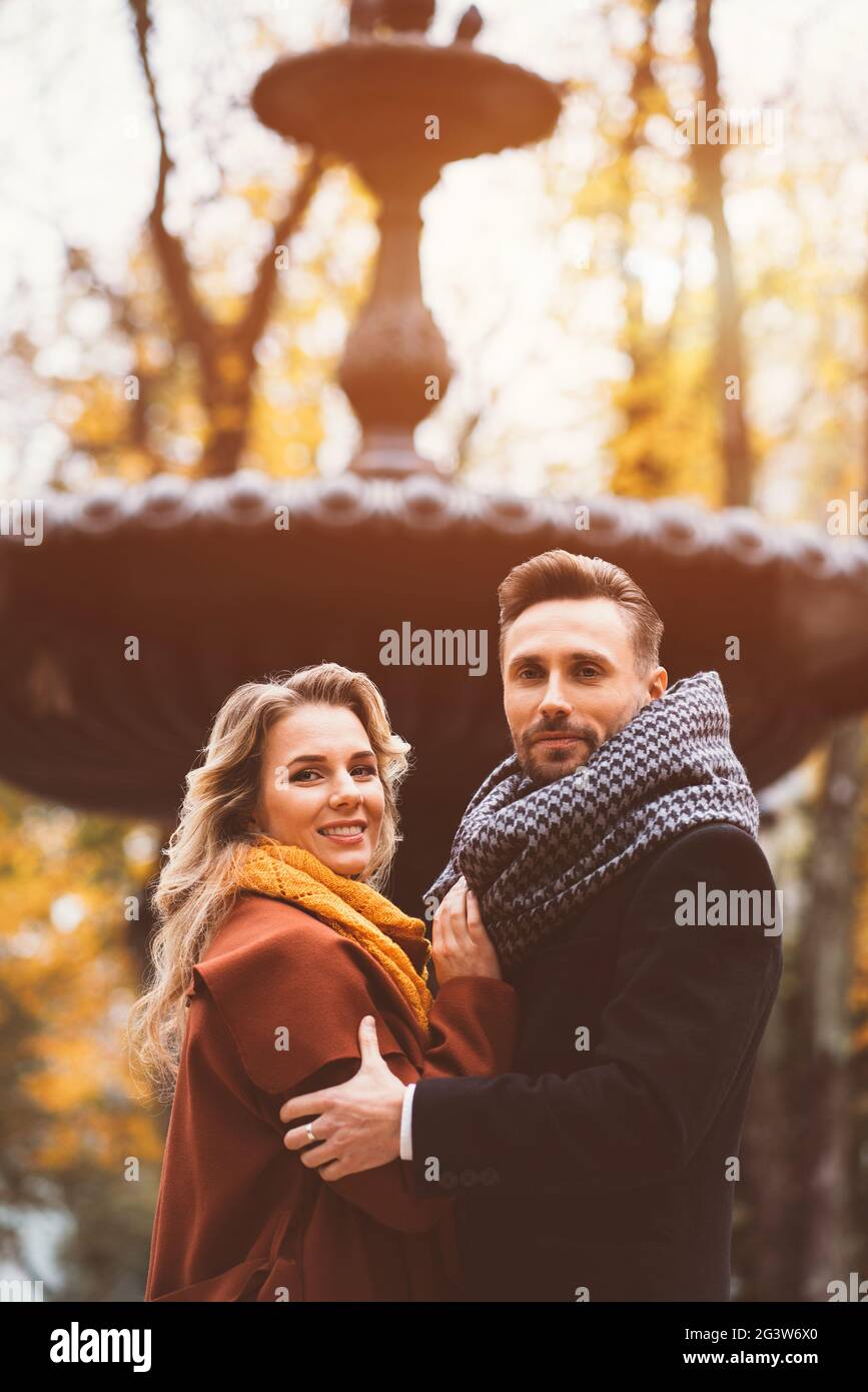 Halblanges Porträt liebevolles Paar junger Menschen, die umarmt stehen und die Kamera mit einem alten Brunnen auf dem Hintergrund A betrachten Stockfoto