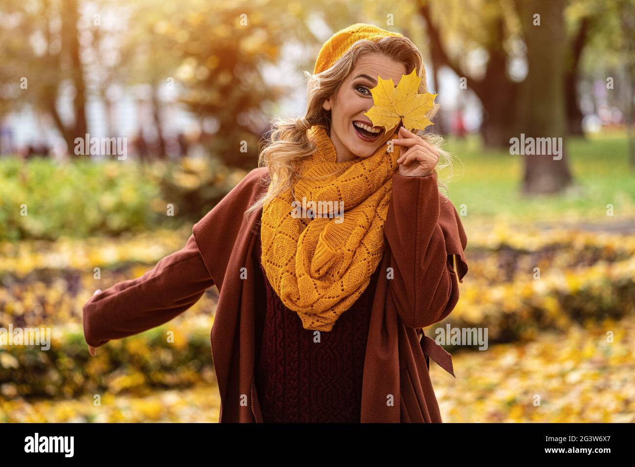 Glücklich lachende Frau verstecken Auge mit einem vergilbten Blatt in gelb gestrickt Baskenmütze mit Herbstblättern in der Hand und Herbst gelben Garten oder Stockfoto