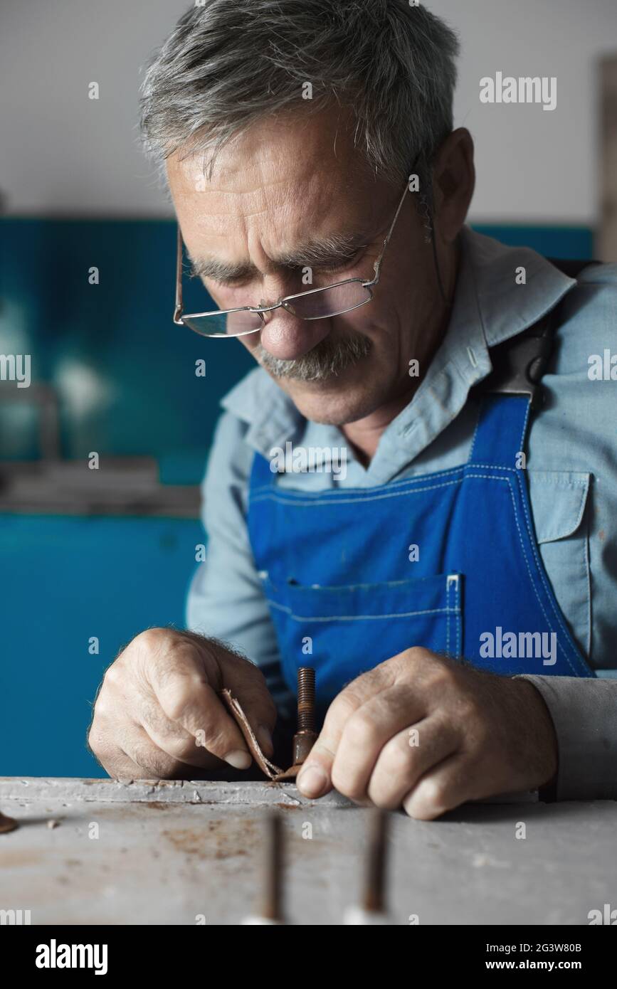 Ein älterer Meister mit Brille bei der Arbeit. Ein weißer alter Mann mit kaukasischem Aussehen sitzt an einem Tisch und arbeitet mit seinen Händen Stockfoto
