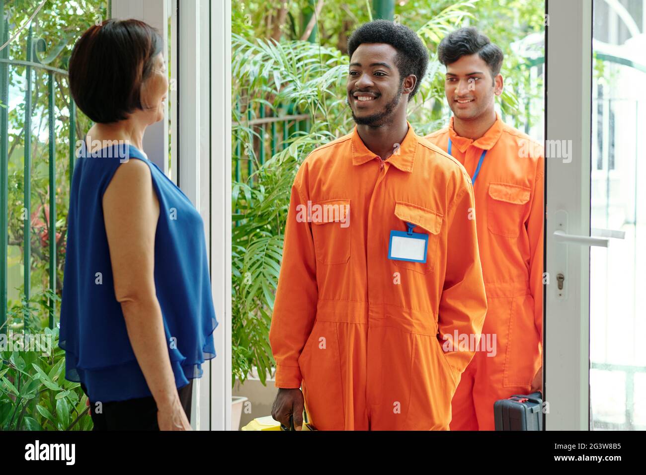 Lächelnde junge, multiethnische Hausmeister in orangefarbenen Bodys stehen am Eingang des Hauses und kommen in das Haus der Kunden, eine reife Frau heißt sie willkommen Stockfoto