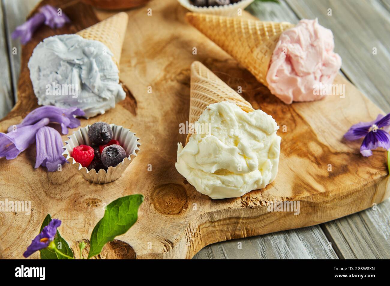 Vanille-, Heidelbeer- und Himbeereis in Waffelkegel auf Holzbrett mit Beeren und Blumen. Stockfoto