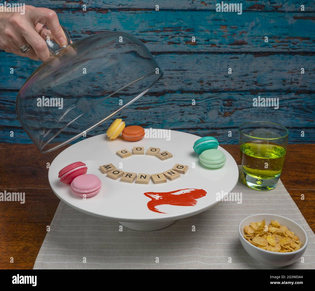 Eine weibliche Hand hebt einen Glasdeckel von einem weißen Kuchenständer, auf dem neben einem Glas Saft und einer Schüssel Müsli steht Stockfoto