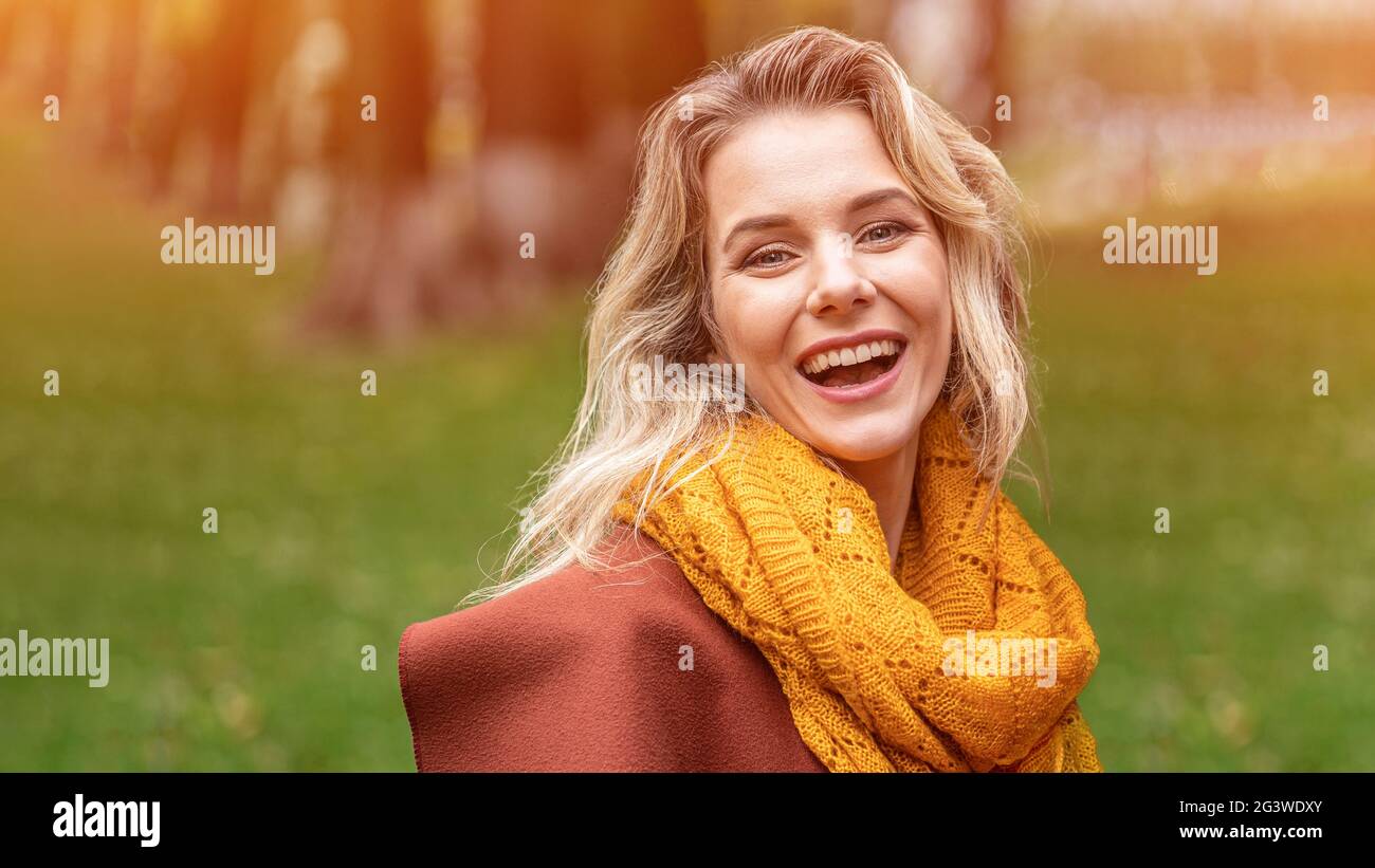 Fröhliche junge Frau im Herbstmantel und gelber Strickschal, die fröhlich lächelnd im herbstgelben Garten oder Park steht. Wunderschön Stockfoto