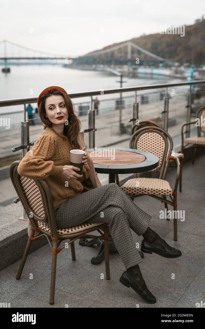 Seitwärts sitzende, junge Pariser Frau auf der Restaurantterrasse. Porträt einer stilvollen jungen Frau in Herbstmantel und rot Stockfoto